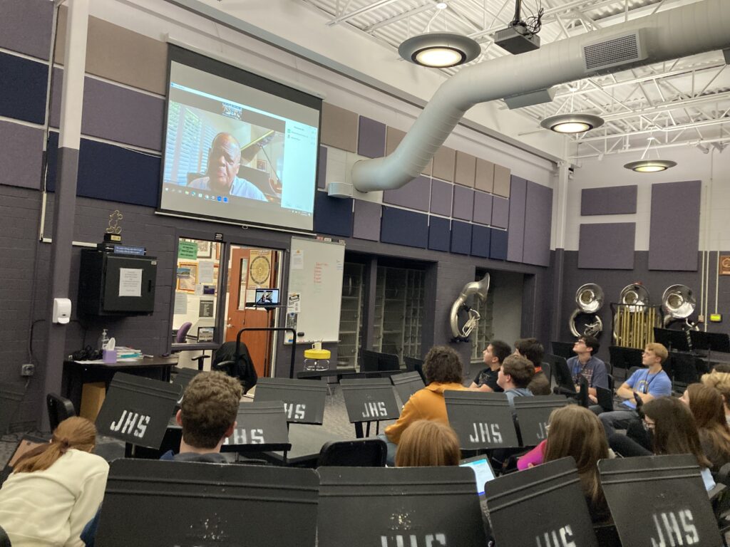 Sylvester Rivers on a projection screen via Zoom. High school music room with tubas along the wall and students watching the screen.